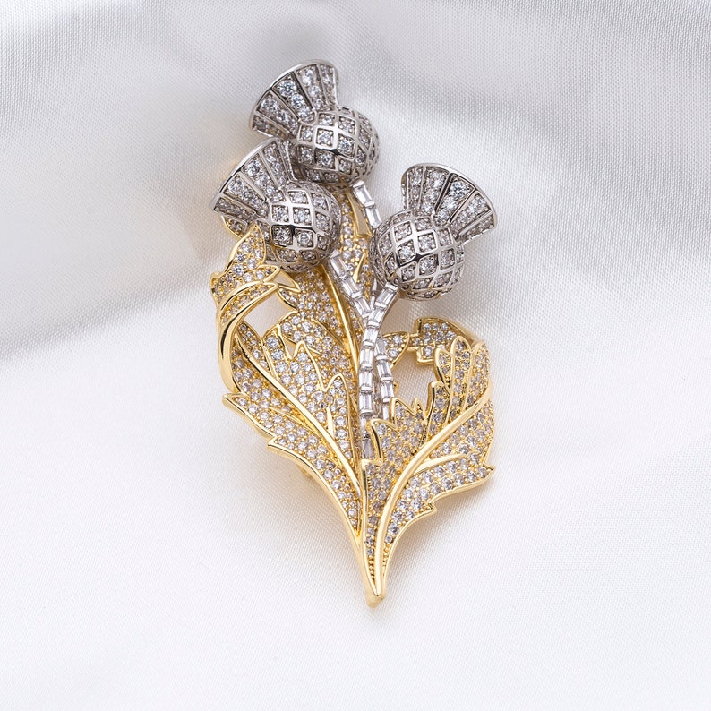 May include: A gold and silver brooch featuring a cluster of three thistle flowers with sparkling rhinestones. The thistle flowers are set against a backdrop of gold leaves, also adorned with rhinestones.