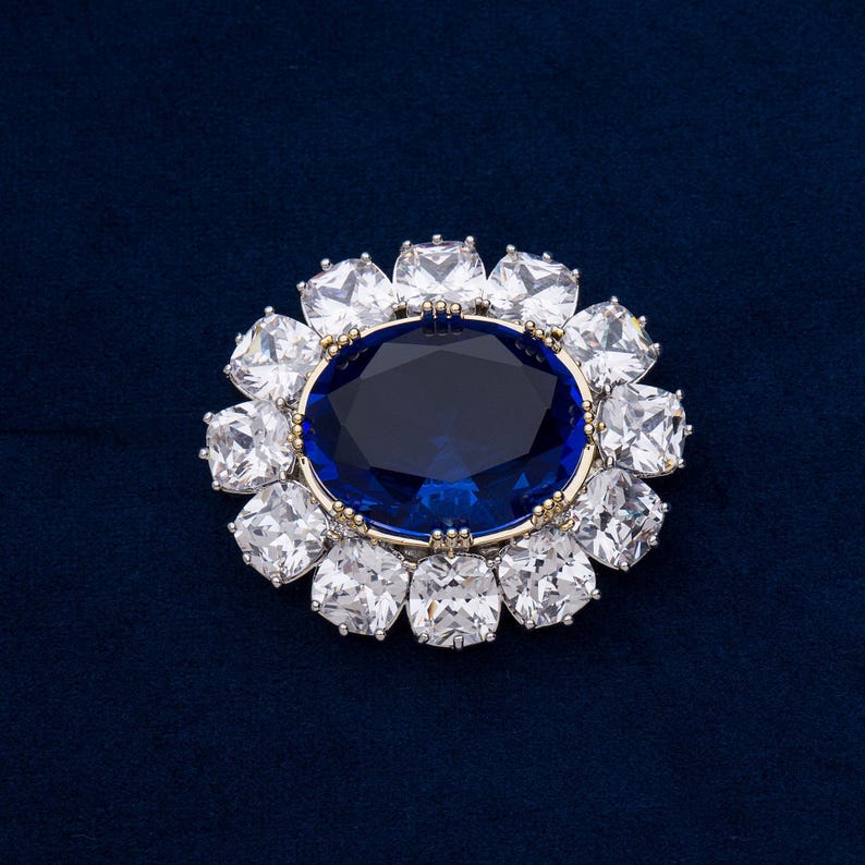 May include: A detailed close-up of a brooch with a large, oval, deep blue gemstone at the center, surrounded by a halo of square, clear gemstones. The setting is gold-toned, and the background is a dark blue fabric.