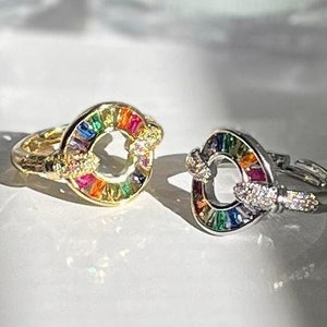 Puede incluir: Dos anillos con un arcoíris de piedras de colores engastadas en un círculo. Un anillo es de oro y el otro de plata.