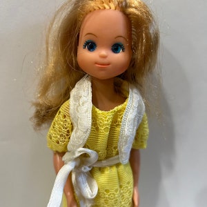 May include: A vintage doll with blonde hair, blue eyes, and a yellow lace dress with a white sash.