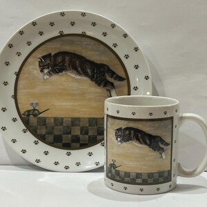 May include: A white ceramic mug and plate set with a brown cat jumping over a mouse. The mug and plate have a brown paw print pattern.
