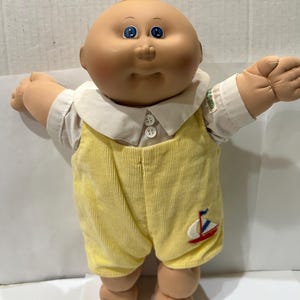 May include: A vintage Cabbage Patch Kid doll with a round face, blue eyes, and a tuft of yellow hair. The doll wears a white collared shirt under yellow corduroy overalls with a sailboat applique. The doll's arms are outstretched.