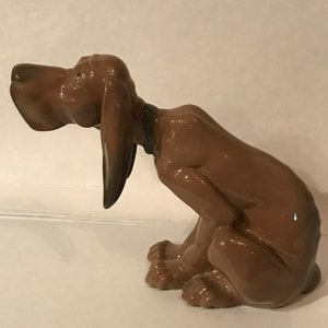 May include: A brown ceramic figurine of a dog sitting with its head tilted down. The dog has a glossy finish and appears to be a bloodhound.