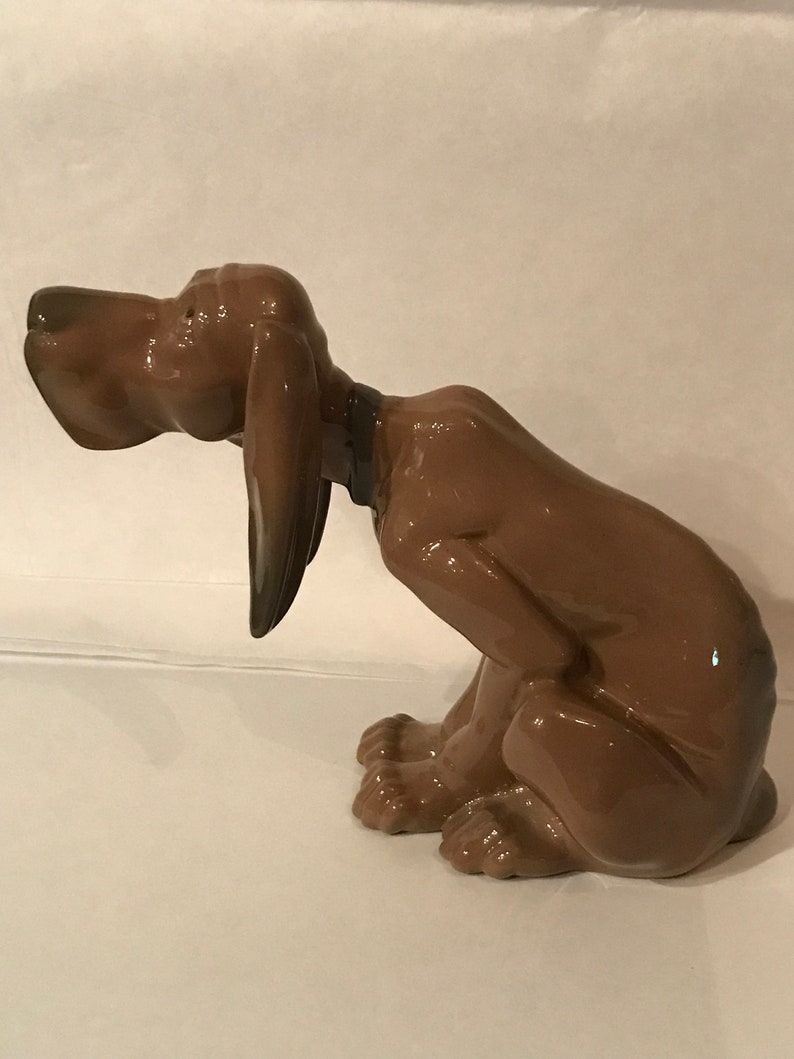 May include: A brown ceramic figurine of a dog sitting with its head tilted down. The dog has a glossy finish and appears to be a bloodhound.