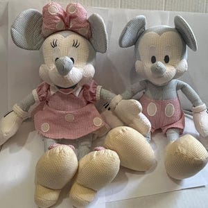 Puede incluir: Dos mu&ntilde;ecos de peluche de los personajes de Disney, Mickey y Minnie Mouse. Minnie lleva un vestido y un lazo a rayas rosas y blancas. Mickey lleva pantalones cortos a rayas rojas y blancas. Ambos tienen orejas y brazos a rayas azules y blancas.