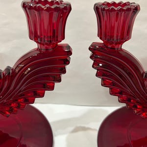 May include: A pair of red glass candle holders with a unique, wave-like design. The holders have a fluted, trumpet-shaped top for candles.