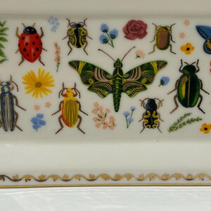 May include: A white rectangular ceramic dish with a gold trim and a colorful insect and flower design. The dish features a large green butterfly, ladybugs, beetles, and flowers.