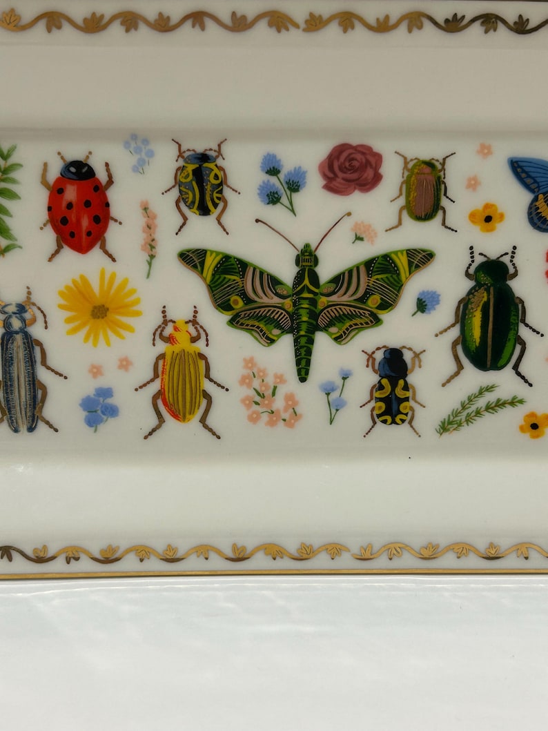 May include: A white rectangular ceramic dish with a gold trim and a colorful insect and flower design. The dish features a large green butterfly, ladybugs, beetles, and flowers.