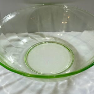 May include: A clear, green-tinted glass mixing bowl with a wide, shallow design. The bowl has a rounded shape with a slightly flared rim and two decorative handles. The base of the bowl is a lighter shade of green. A vintage kitchenware item.