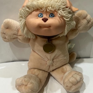 May include: A vintage plush toy in the shape of a dog with a light tan body and a cream-colored, shaggy mane. The toy has blue eyes, a brown nose, and a brown collar with a gold-colored medallion. The dog is in a sitting position with its arms raised.