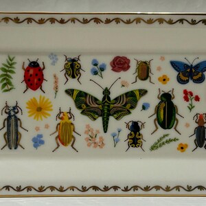 May include: A white rectangular ceramic dish with a gold trim and a colorful insect and butterfly design. The dish features a variety of insects, including ladybugs, butterflies, and beetles, all with intricate details.