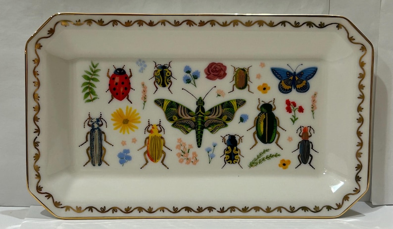 May include: A white rectangular ceramic dish with a gold trim and a colorful insect and butterfly design. The dish features a variety of insects, including ladybugs, butterflies, and beetles, all with intricate details.
