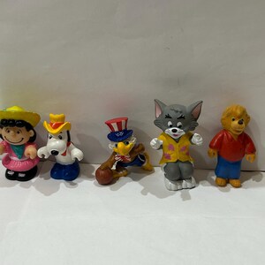 May include: A set of five small plastic figurines. The figurines are of various cartoon characters, including a girl in a pink dress, a dog in a blue and white uniform, an eagle in a red, white, and blue uniform, a cat in a yellow shirt, and a bear in a red shirt and blue pants.