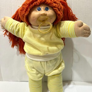 May include: A vintage Cabbage Patch Kid doll with orange yarn hair, blue eyes, and a pacifier. The doll is wearing a yellow long-sleeved shirt and matching shorts with yellow and white striped accents. The doll's arms are outstretched.