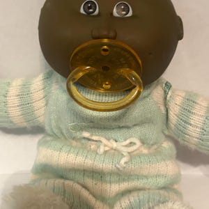 May include: A doll with a dark complexion, brown eyes, and a round head. The doll has a translucent amber-colored plastic pacifier in its mouth and is wearing a light mint green and white horizontally striped knitted long-sleeve onesie.