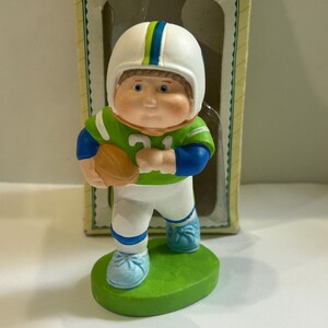 May include: A ceramic figurine of a football player in a green and blue uniform, white helmet, and holding a football. The figurine is standing on a green oval base. The figurine is in front of a box with a window.