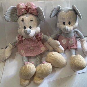 Puede incluir: Dos mu&ntilde;ecos de peluche de personajes de Disney. Uno es Minnie Mouse, con un vestido y lazo a rayas rosas y blancas. El otro es Mickey Mouse, con pantalones cortos a rayas rojas y blancas. Ambos tienen pies y manos color crema.