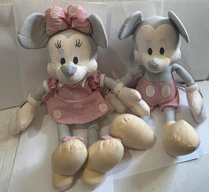Puede incluir: Dos mu&ntilde;ecos de peluche de personajes de Disney. Uno es Minnie Mouse, con un vestido y lazo a rayas rosas y blancas. El otro es Mickey Mouse, con pantalones cortos a rayas rojas y blancas. Ambos tienen pies y manos color crema.