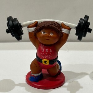 May include: A small plastic figurine of a weightlifter wearing a red, white, and blue USA Olympic uniform. The figure is lifting a barbell with black weights on each end.