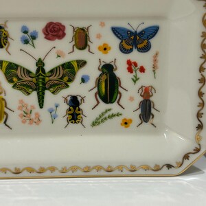 May include: A white rectangular ceramic dish with a gold trim and a decorative design of colorful butterflies and beetles. The dish is perfect for serving snacks or holding jewelry.