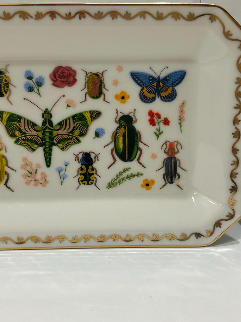 May include: A white rectangular ceramic dish with a gold trim and a decorative design of colorful butterflies and beetles. The dish is perfect for serving snacks or holding jewelry.