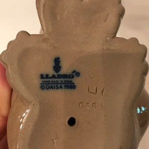 May include: A beige ceramic figurine of a woman holding a bird. The figurine is marked with the Lladro logo, "Hand Made in Spain", and "Daisa 1980". The figurine is also marked with the number "6588".