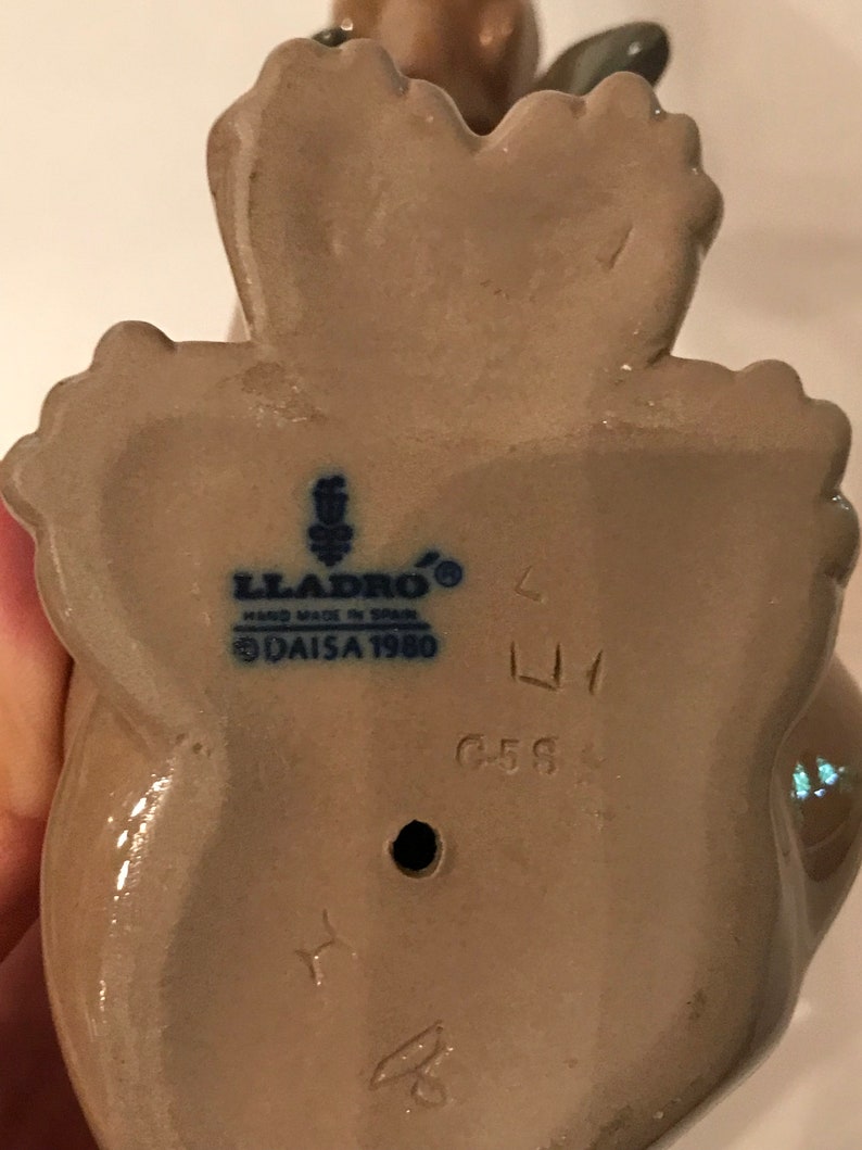 May include: A beige ceramic figurine of a woman holding a bird. The figurine is marked with the Lladro logo, "Hand Made in Spain", and "Daisa 1980". The figurine is also marked with the number "6588".