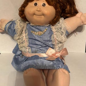 May include: A doll with auburn hair tied in a ponytail with a purple scrunchie, featuring a round face and brown eyes. The doll wears a blue satin dress with white lace trim, a small floral embroidery on the chest, and a light pink ribbon bow at the waist.