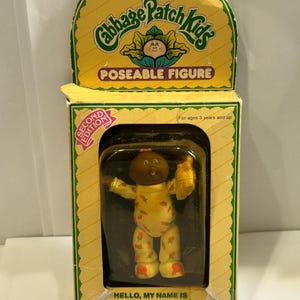 May include: A vintage Cabbage Patch Kids poseable figure in its original packaging. The doll is wearing a yellow onesie with strawberry accents and is holding a yellow accessory. The box is yellow with green accents and features the text "POSEABLE FIGURE".