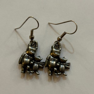 May include: A pair of silver tone dangle earrings featuring Winnie the Pooh and Piglet. The characters are holding hands and are standing on a small base.