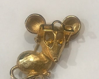 May include: A gold-toned mouse brooch with yellow rhinestone eyes. The mouse is wearing a gold-toned frame around its eyes.