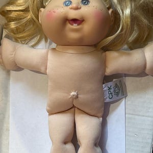 May include: A vintage Cabbage Patch Kid doll with blonde hair styled in pigtails. The doll has blue eyes, rosy cheeks, and a smiling expression. The doll's body is made of soft fabric, and a tag with the letters "CPK" is attached.