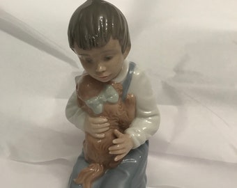 May include: A porcelain figurine of a boy kneeling and holding a small dog. The boy is wearing blue overalls and a white shirt. The dog is brown and has a blue bow.