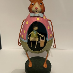 May include: A standing rabbit figurine with a textured orange head and long ears, wearing a light blue bow tie. The body is a pink egg shape with yellow polka dots, featuring a hollow center containing a smaller rabbit holding a woven basket.