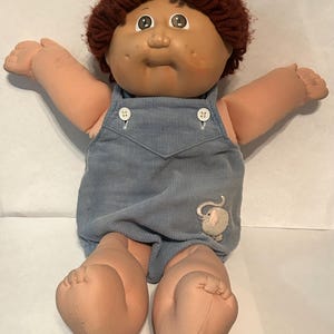 May include: A doll with a round face, tan skin, and short, curly dark brown yarn hair. The doll wears blue corduroy overalls featuring two white buttons at the chest and a small, embroidered light gray elephant patch on the lower left side.