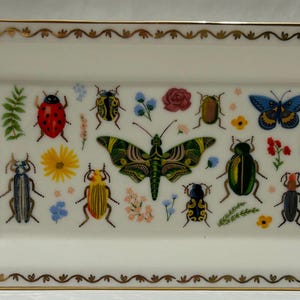 May include: A white rectangular ceramic tray with a gold trim and a design of various insects and flowers. The insects include butterflies, ladybugs, and beetles in shades of red, blue, green, and yellow.