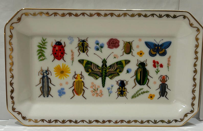 May include: A white rectangular ceramic tray with a gold trim and a design of various insects and flowers. The insects include butterflies, ladybugs, and beetles in shades of red, blue, green, and yellow.
