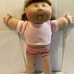 May include: A Cabbage Patch Kids doll with brown hair, blue eyes, and rosy cheeks. The doll is wearing a pink sweater, striped shorts, and a white headband. The doll's arms are outstretched.