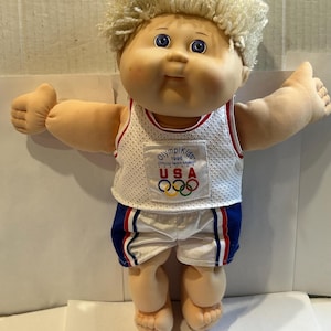 May include: A Cabbage Patch Kids doll wearing a white and red USA Olympic team uniform. The doll has blonde, curly hair, blue eyes, and is giving a thumbs-up gesture with both hands. The uniform includes a tank top and shorts.