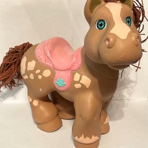 May include: A brown plastic toy horse with a white blaze on its face, cream-colored spots on its body, and a pink saddle. The horse features teal-colored eyes, brown yarn hair for the mane and tail, and a small teal flower detail on the saddle.