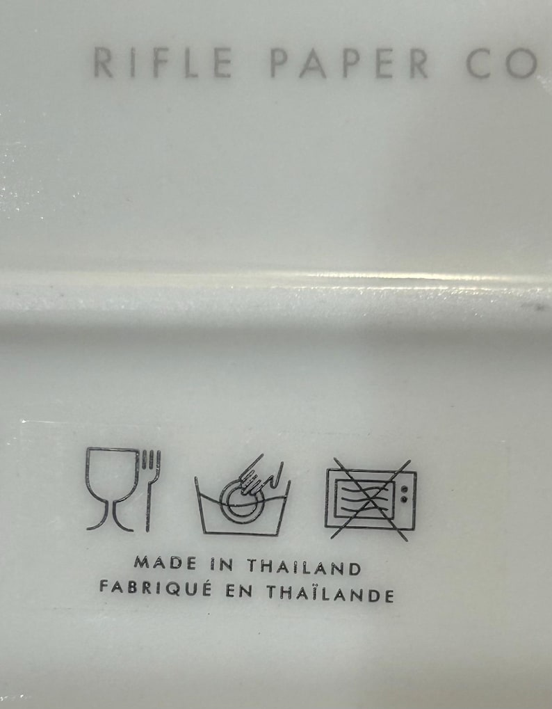 May include: A white ceramic plate with black symbols indicating it is dishwasher and microwave safe. The plate also has the text "RIFLE PAPER CO" and "MADE IN THAILAND FABRIQU&Eacute; EN THA&Iuml;LANDE".