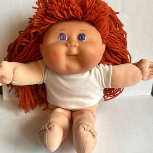 May include: A doll with a round face, light skin, and purple eyes. The doll has thick, textured orange yarn hair and is wearing a plain white short-sleeved shirt. The doll has a soft, fabric body with visible stitching on the feet and hands.