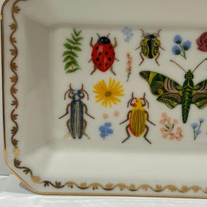 May include: A white ceramic rectangular tray with a gold trim and a decorative design of colorful insects and flowers. The tray features a ladybug, a butterfly, and several beetles.