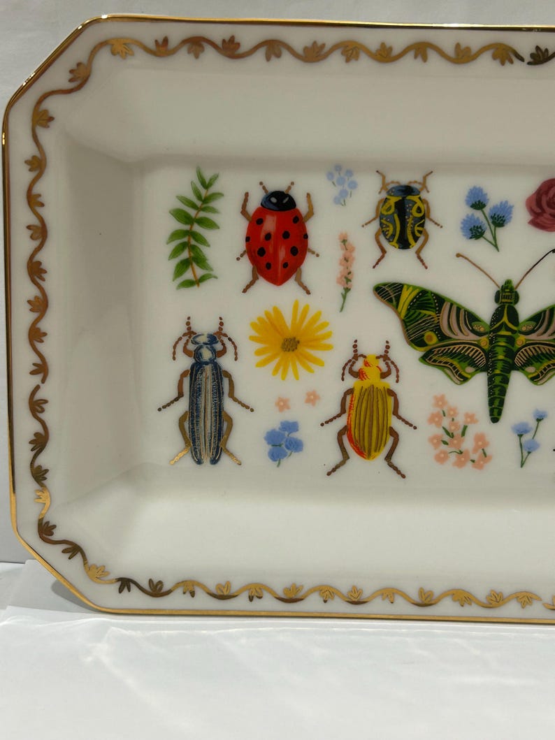 May include: A white ceramic rectangular tray with a gold trim and a decorative design of colorful insects and flowers. The tray features a ladybug, a butterfly, and several beetles.