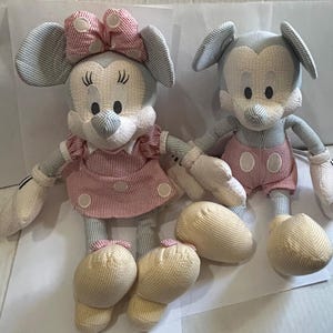 Puede incluir: Dos mu&ntilde;ecos de peluche de personajes de dibujos animados. Una mu&ntilde;eca lleva un vestido y un lazo a rayas rosas y blancos. La otra mu&ntilde;eca lleva pantalones cortos rosas. Ambas mu&ntilde;ecas tienen orejas grandes, guantes blancos y zapatos amarillos. Las mu&ntilde;ecas est&aacute;n hechas de tela suave y texturizada.