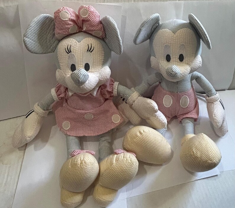 Puede incluir: Dos mu&ntilde;ecos de peluche de personajes de dibujos animados. Una mu&ntilde;eca lleva un vestido y un lazo a rayas rosas y blancos. La otra mu&ntilde;eca lleva pantalones cortos rosas. Ambas mu&ntilde;ecas tienen orejas grandes, guantes blancos y zapatos amarillos. Las mu&ntilde;ecas est&aacute;n hechas de tela suave y texturizada.