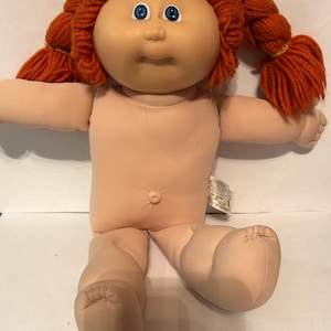 May include: A doll with a soft fabric body and a molded vinyl head featuring bright blue eyes, a small nose, and a closed mouth. The doll has vibrant orange yarn hair styled into two pigtails tied with orange bands and a visible belly button.