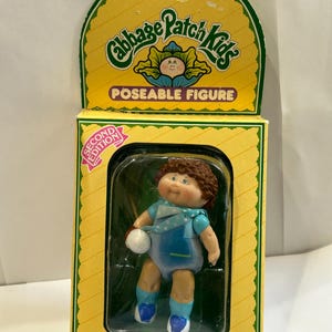 May include: A Cabbage Patch Kids poseable figure in its original packaging. The doll has brown curly hair, blue overalls, and blue shoes. The packaging is yellow and green with the words "Cabbage Patch Kids" and "Poseable Figure". The doll's name is John Davis.