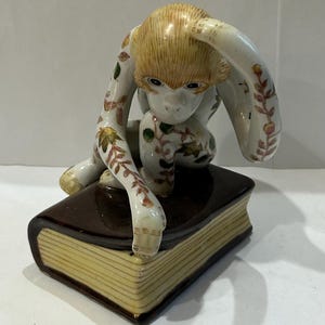 May include: A ceramic monkey figurine with a cream-colored body, sitting atop a brown book. The monkey has a light brown head and is decorated with floral patterns in green, yellow, and red. The book has yellowed pages.