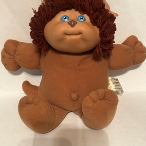 May include: A brown plush doll with a soft fabric body, bright blue eyes, and a dark brown yarn hairstyle. The doll features a small nose, a simple stitched mouth, and a small tail. A white fabric tag is attached to the side of the doll's torso.